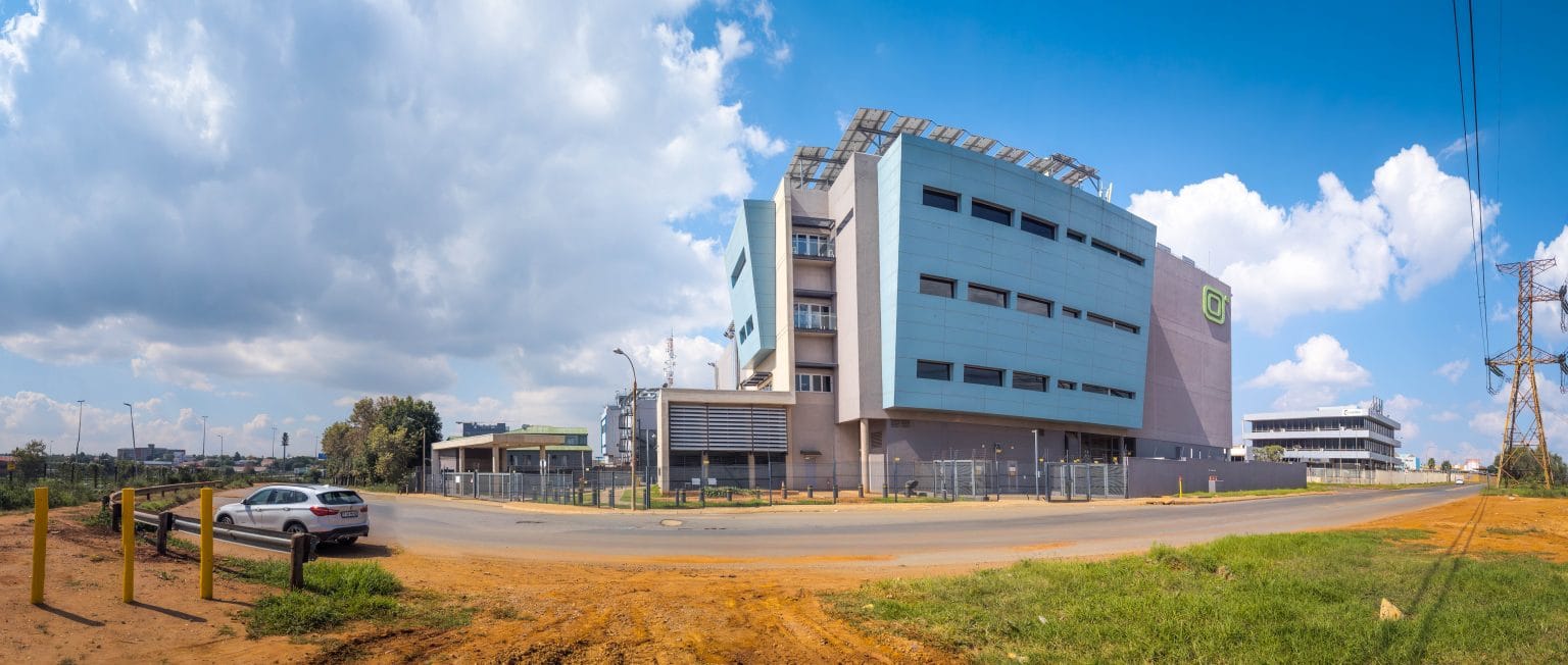 Vista amplia de un edificio de centro de datos con paneles azules y grises y placas solares en el tejado, en un día soleado en el campus de Isando de Teraco.