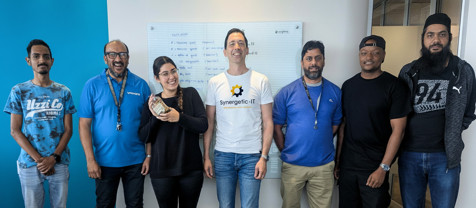 Group of seven people standing side by side against a blue and white office wall with a whiteboard behind them, posing for a team photo, with their faces intentionally blurred for privacy.