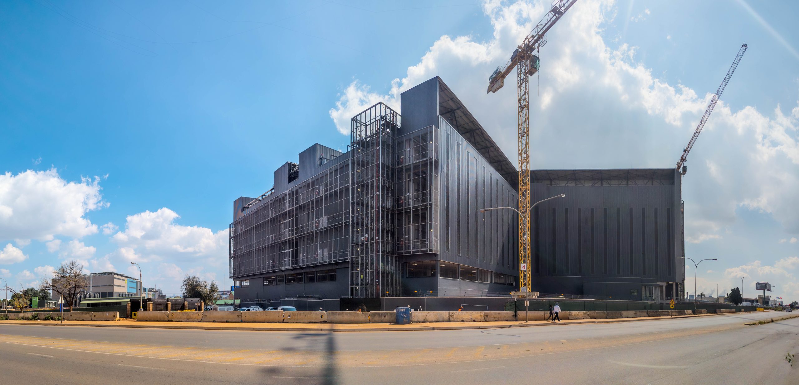 A modern data center building with light blue and gray exterior panels stands beside a quiet road under a partly cloudy blue sky, with a parked car in the foreground, security fencing around the facility, and power lines and another office building visible in the distance.