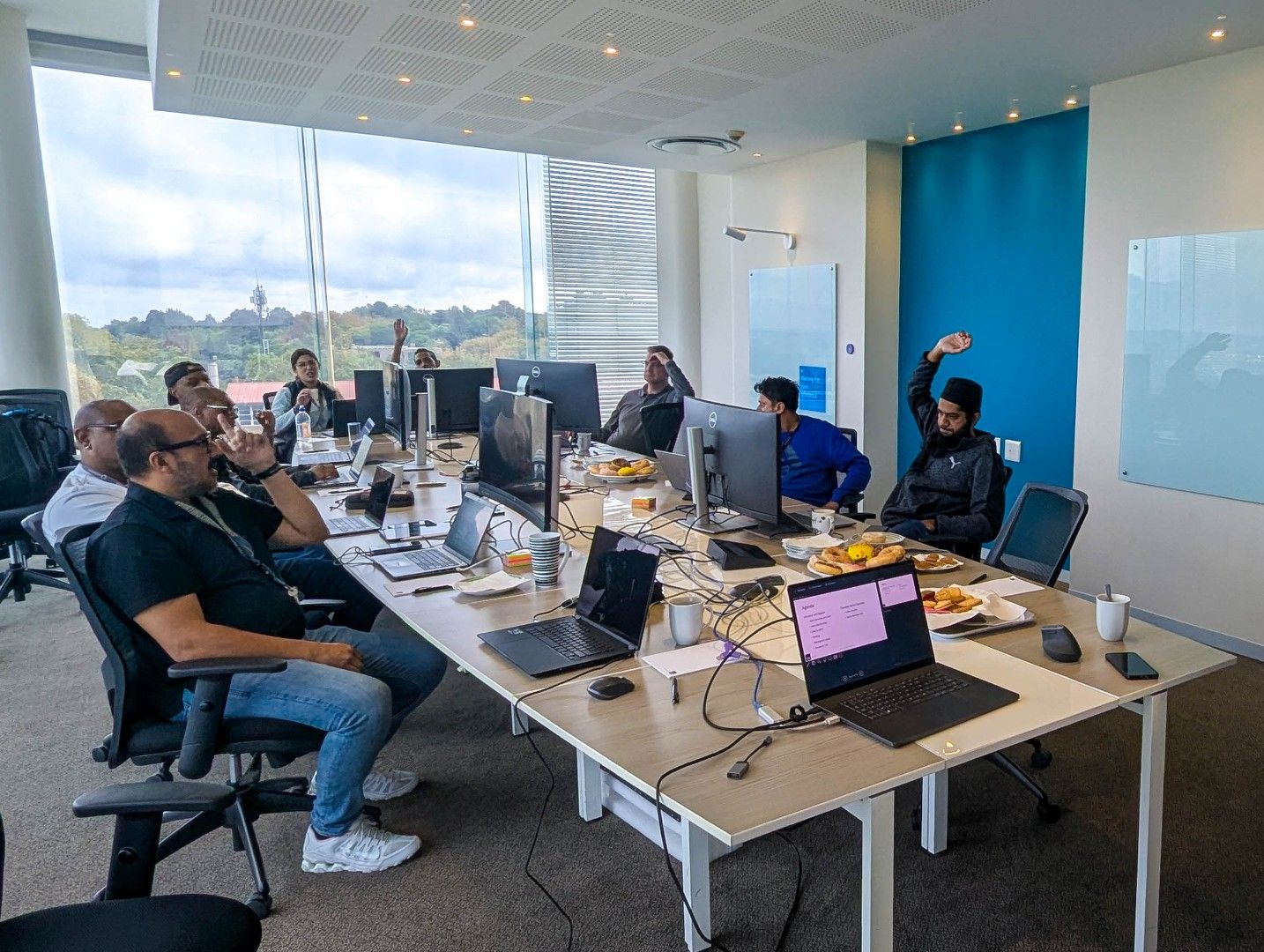 Group of people in a modern office meeting room sitting around a large table with laptops, monitors, cables, and snacks, participating in a collaborative training or workshop while facing a wall of windows with a city and tree-lined view.
