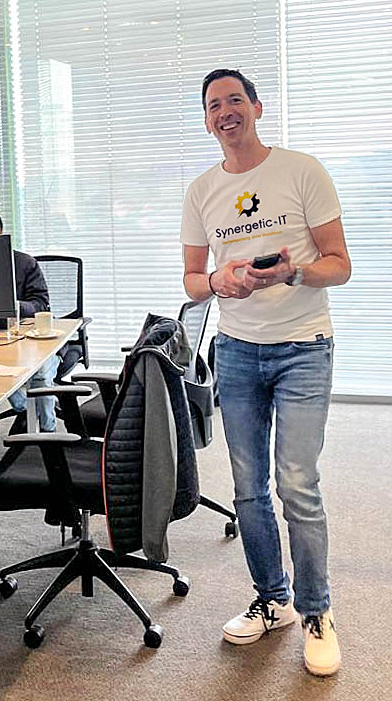 A blurred participant in an office wearing a white Synergetic IT t-shirt, walking between desks with laptops and chairs in a bright room with large window blinds.